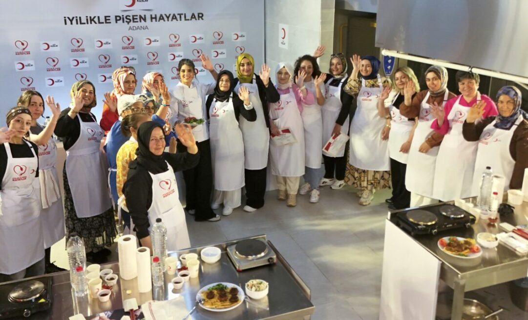 Group of women in white aprons wave at the camera during a cooking charity event in Adana, smiling in a kitchen studio background with a banner behind them.