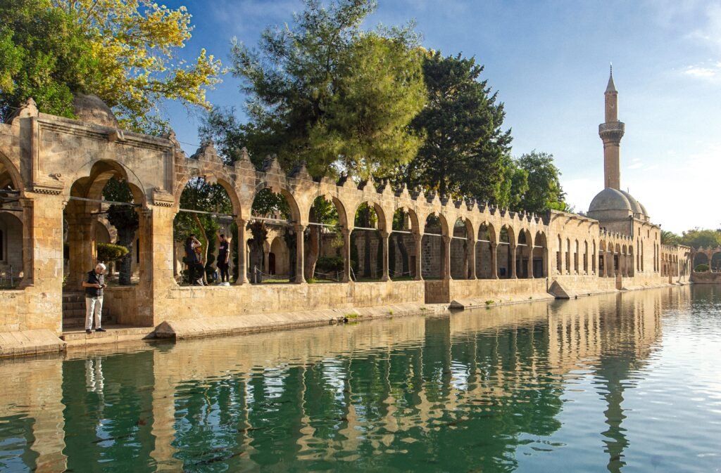 Stone arched colonnade along a calm waterway, with trees behind and a tall mosque minaret in the background.