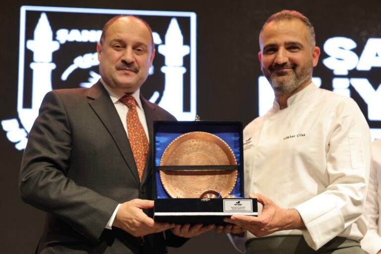 Two men in formal attire presenting and receiving a round copper plate award in a blue presentation case on a stage with a backdrop of logos.