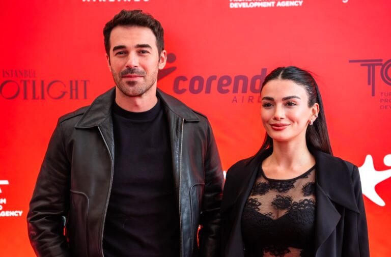 Man and woman posing on a red carpet in front of a red backdrop with event logos.