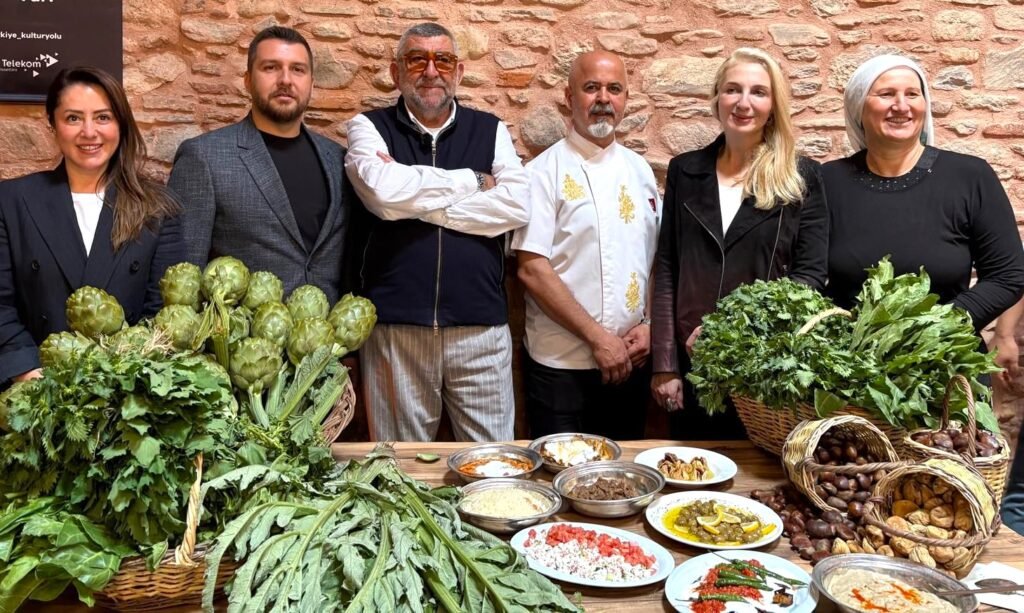 Six people pose behind a table laden with artichokes, leafy greens, baskets of root vegetables, and plates of prepared dishes against a brick wall.