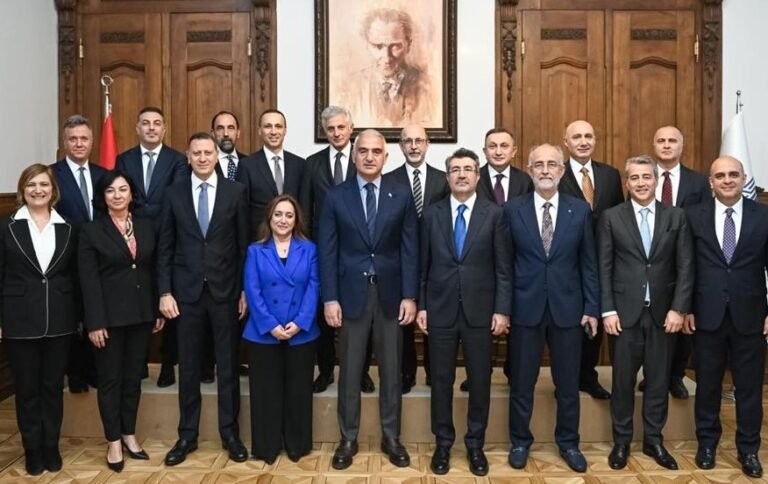 Group of government officials in formal attire posing for a portrait in a wood-paneled room, with a framed portrait and national flags in the background.