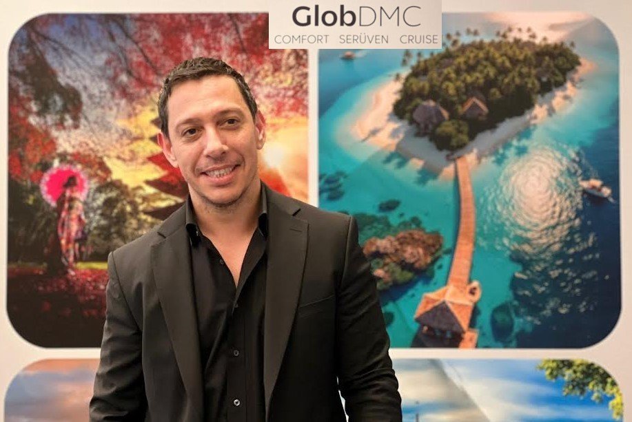 Smiling man in a black suit stands in front of travel posters at a GlobDMC display showing tropical island and autumn scenery nearby.