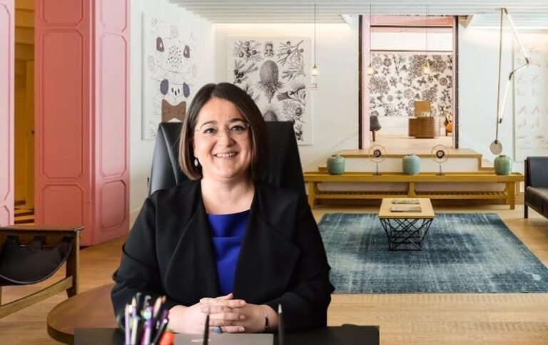 Smiling woman in a black blazer and blue blouse sits at a desk in a bright, stylish reception area with art on the walls and a blue rug in the background.