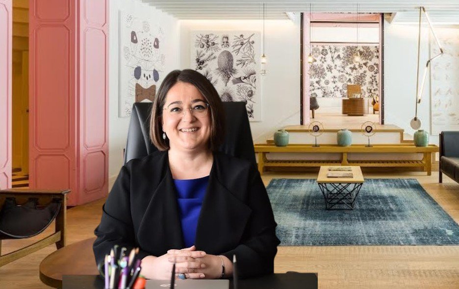 Smiling woman in a black blazer and blue blouse sits at a desk in a bright, stylish reception area with art on the walls and a blue rug in the background.