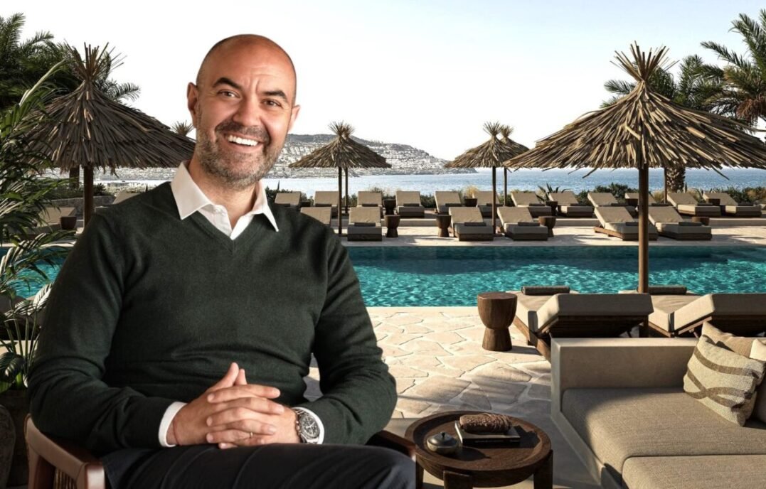 Smiling man in a dark green sweater seated at a resort pool patio with thatch umbrellas and lounge chairs nearby, ocean in the background.