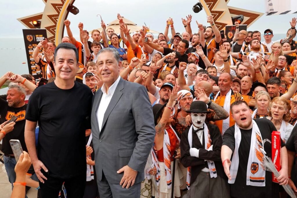 Two men pose for a photo at a crowded beach‑side fan event, surrounded by hundreds of orange‑clad supporters waving their arms.