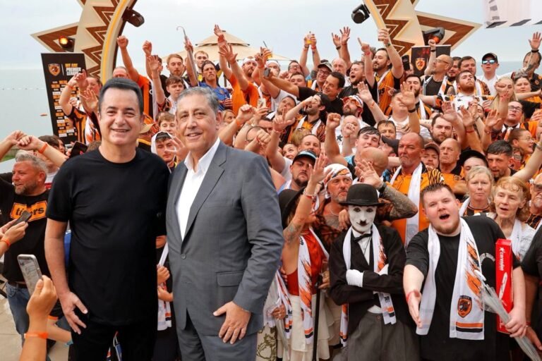 Two men pose for a photo at a crowded beach‑side fan event, surrounded by hundreds of orange‑clad supporters waving their arms.