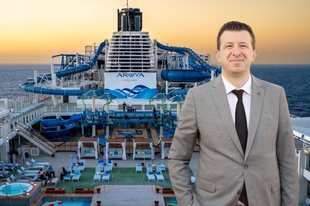 Man in a gray suit and tie stands on a cruise ship deck at sunset, with blue water slides and a pool behind him.