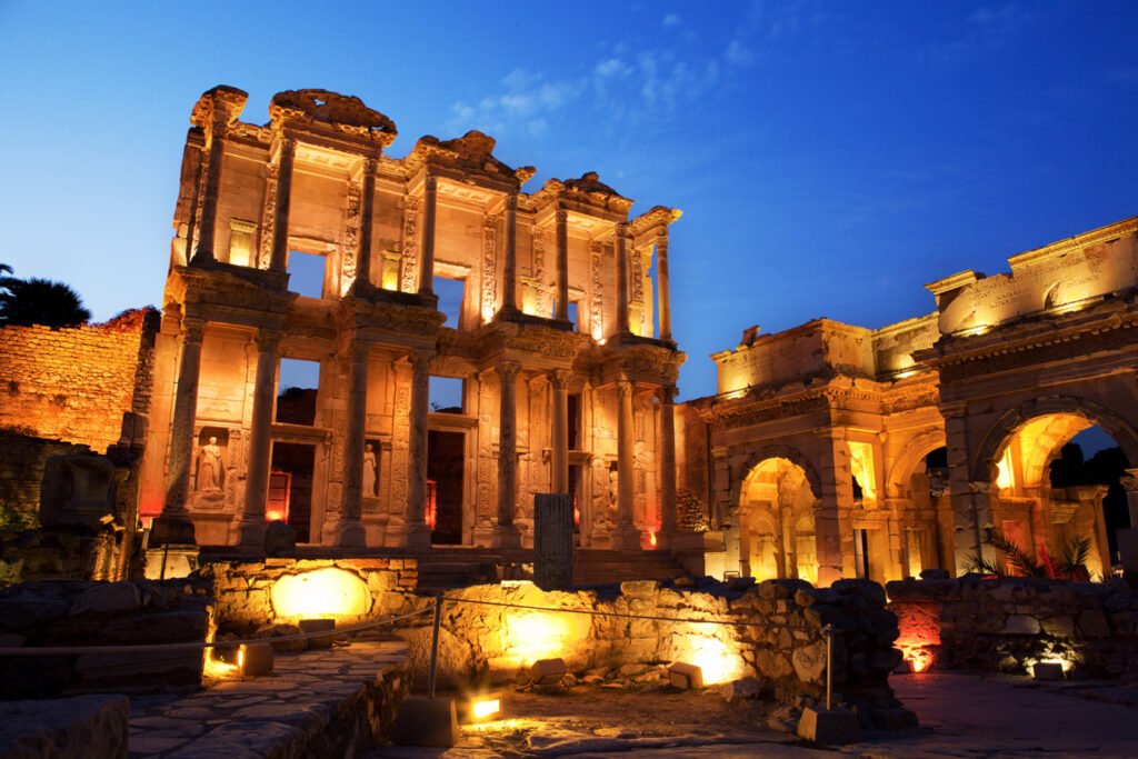 Night view of ancient stone ruins with tall, illuminated columns and arches under a blue sky