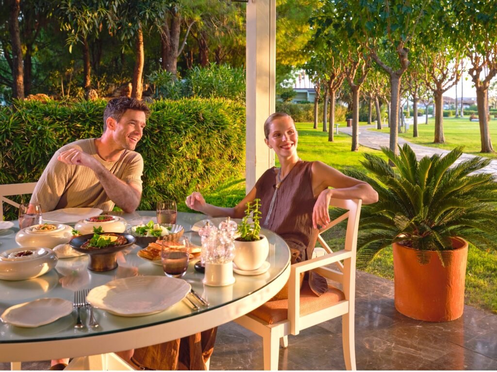 Couple enjoying an outdoor meal at a glass-topped table on a patio, with food and drinks in view and a garden backdrop.