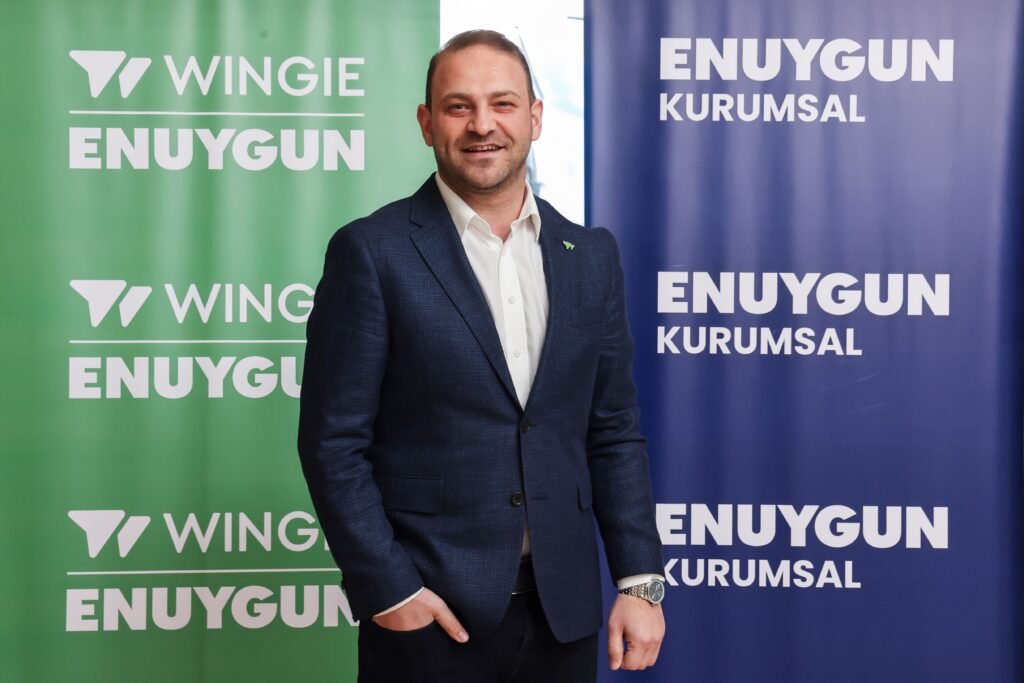 Smiling man in a navy suit and white shirt stands between green and blue banners with ENUYGUN text behind him.