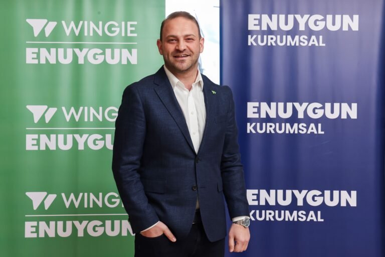 Smiling man in a navy suit and white shirt stands between green and blue banners with ENUYGUN text behind him.
