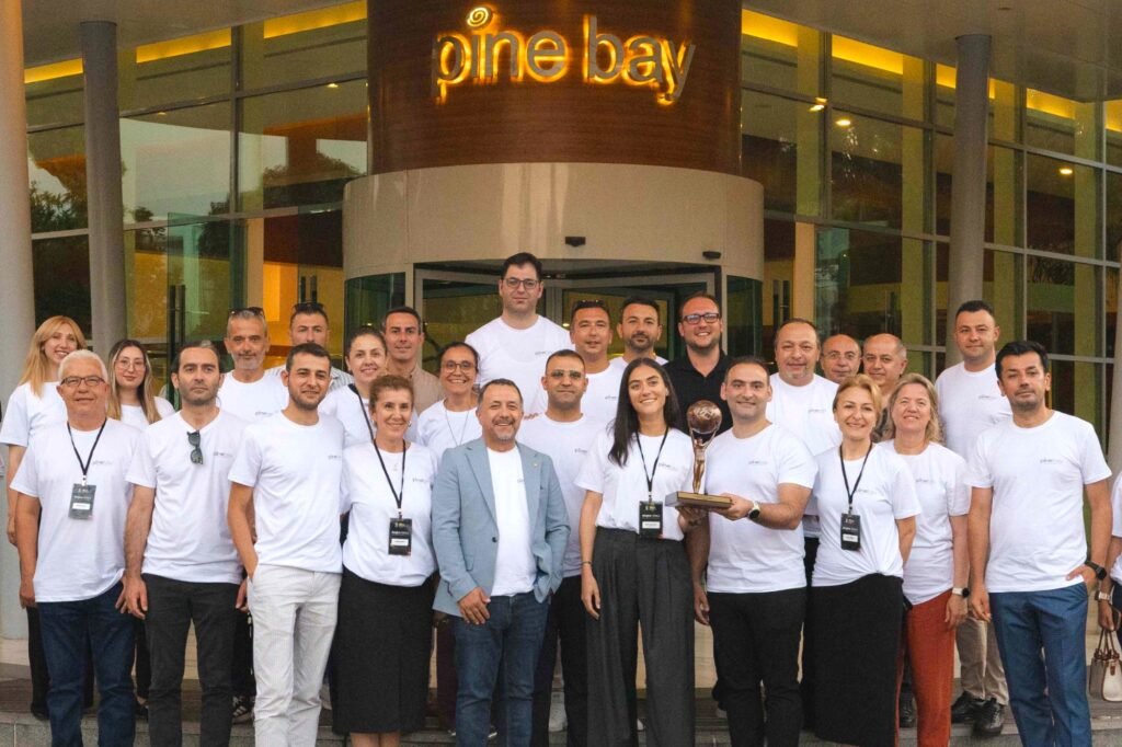 Group of around 20 people in white shirts pose together on the steps outside Pine Bay, with a person holding a trophy in the center.
