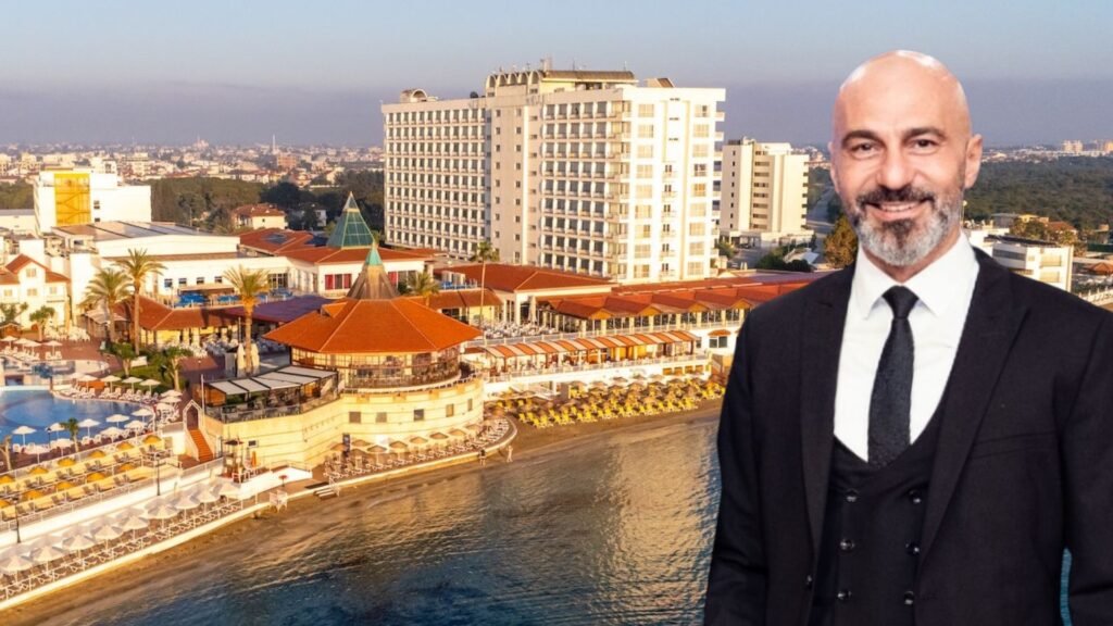 Smiling bald man in a black suit stands in front of a seaside hotel and resort complex with orange-roofed buildings by the water.