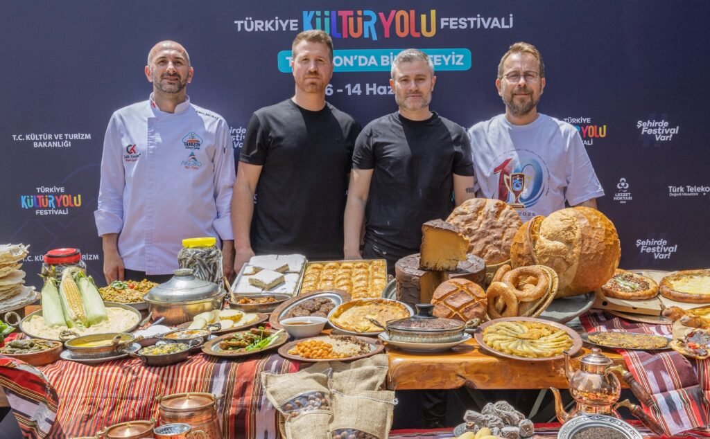 Four men stand behind a long table overflowing with breads and traditional dishes at Türkiye Kültür Yolu Festival.