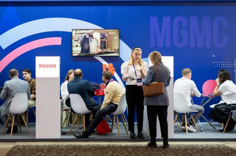 Trade show booth with a blue backdrop and 'MGMC' lettering; people sit at tables and chat, while two women stand and talk nearby.