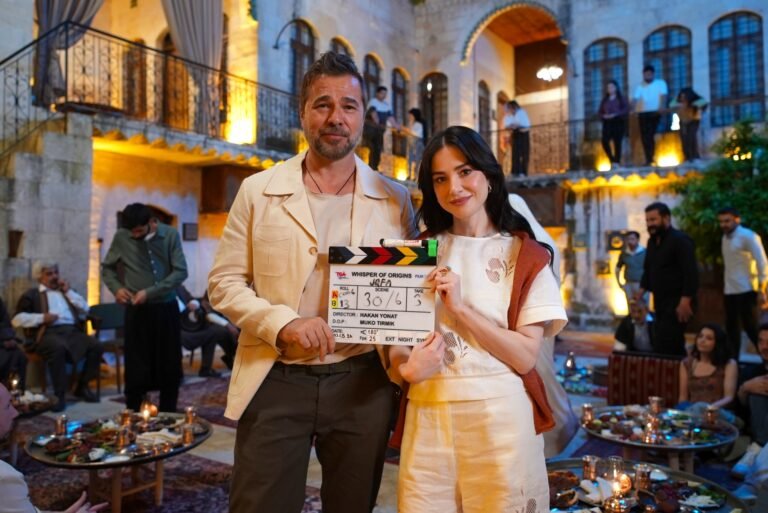 Couple posing with a film clapperboard at a festive outdoor courtyard gathering; warm lights and guests in the background, tables with food nearby.