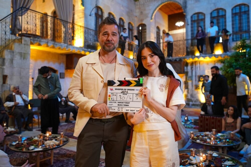 Couple posing with a film clapperboard at a festive outdoor courtyard gathering; warm lights and guests in the background, tables with food nearby.