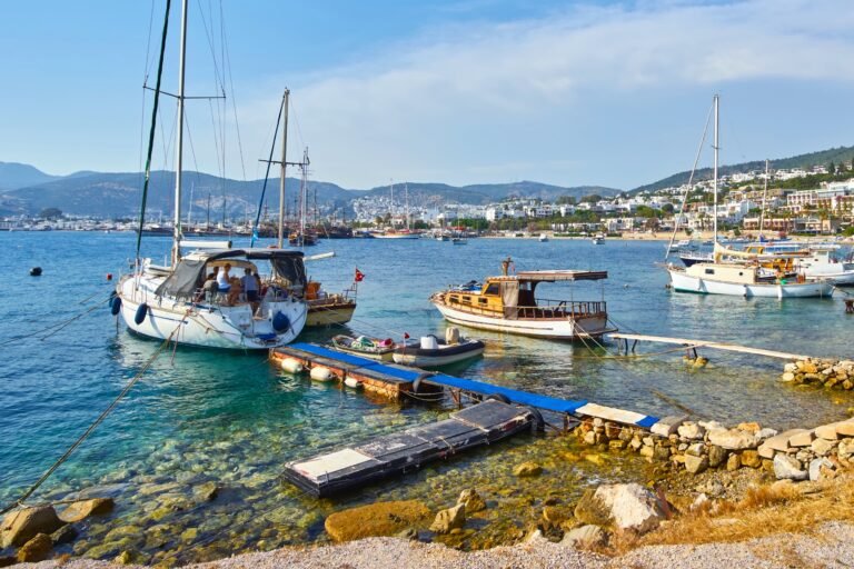 Harbor scene with several sailboats and a wooden boat tied to a floating dock near a rocky shore, a town in the distance.