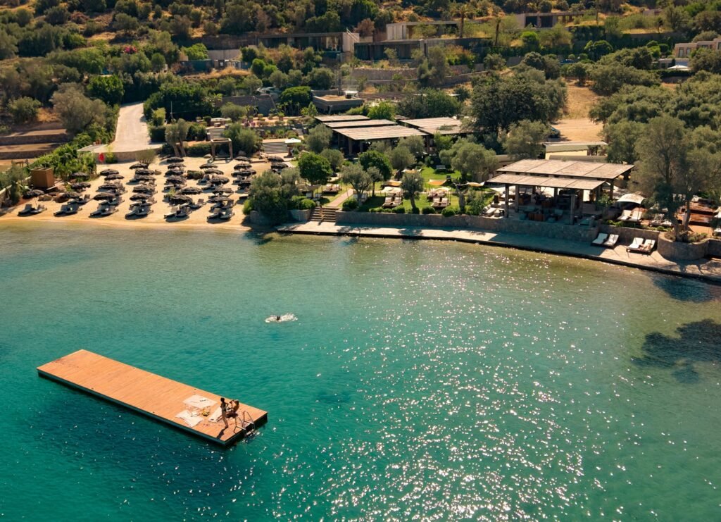 Aerial view of a seaside resort: a long wooden dock extends into turquoise water, with lounge chairs lining the sandy beach and shaded pavilions among green trees.