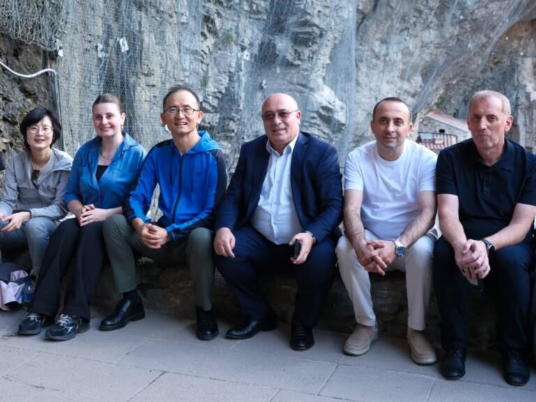 Six people sit side by side on a stone ledge outdoors, smiling at the camera against a rocky wall backdrop.