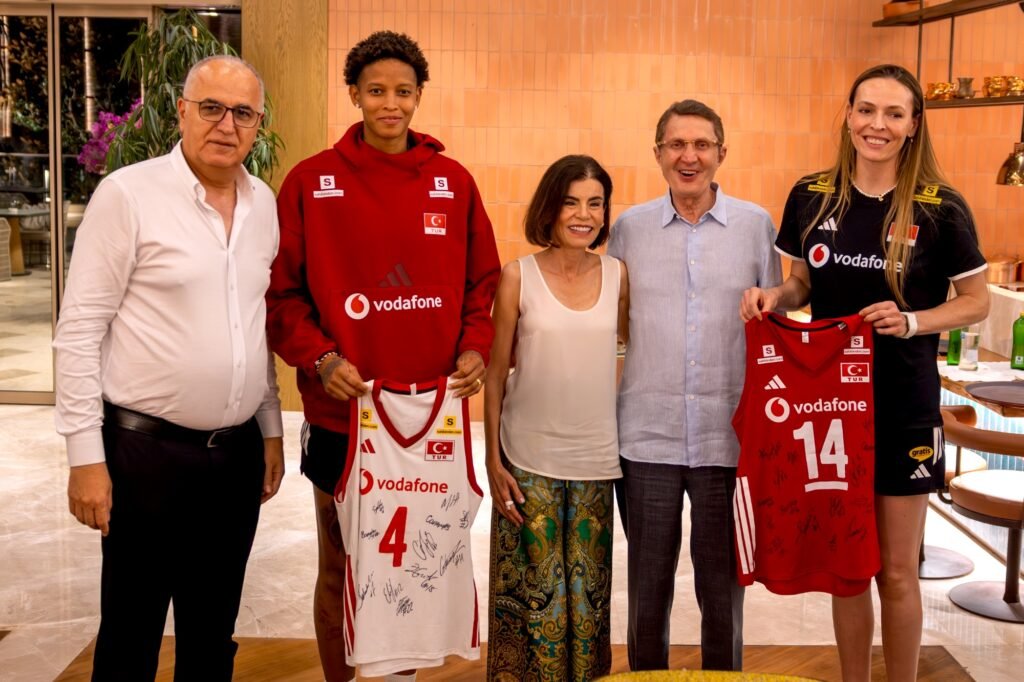 Five people pose indoors as two athletes hold signed red Vodafone basketball jerseys (numbers 4 and 14) with signatures visible on the fabric, in a bright lobby setting.