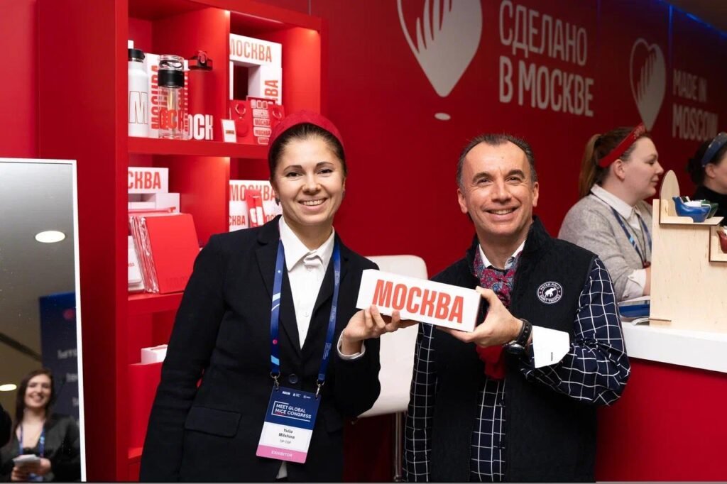 Two smiling event attendants pose in a red booth, one holding a sign that reads МОСКВА (Moscow). The scene shows a Moscow-themed display with products on red shelves in the background.