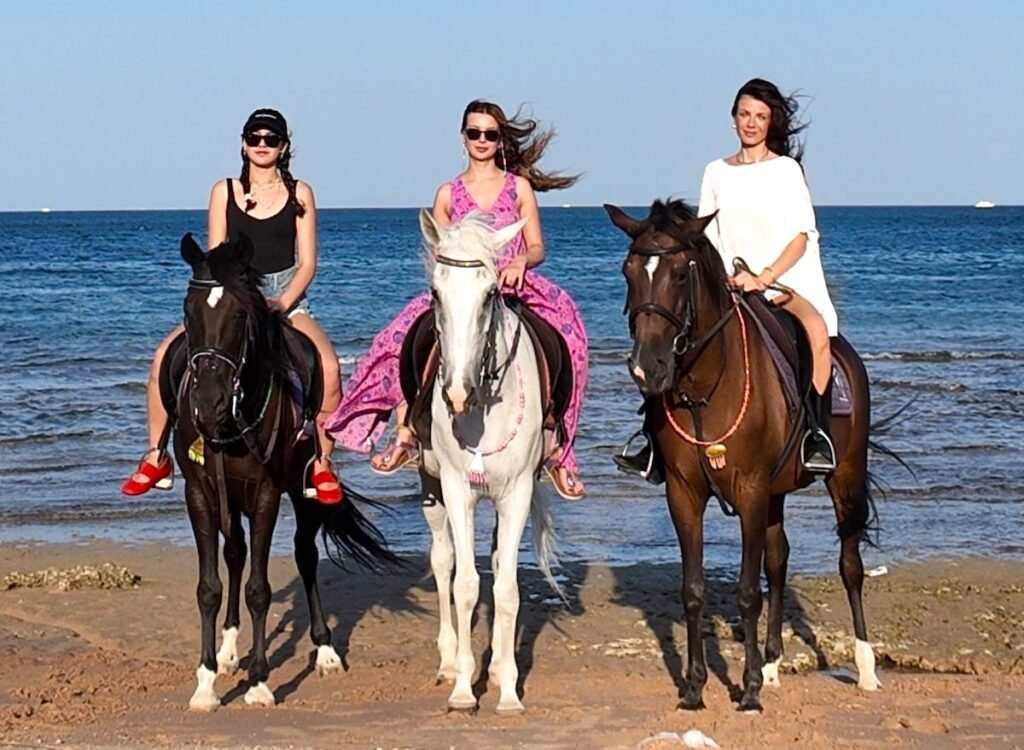 Three women ride horses along a sandy beach with the blue sea behind them; left rider wears black top and red sandals, center wears a pink dress, right wears a white dress.
