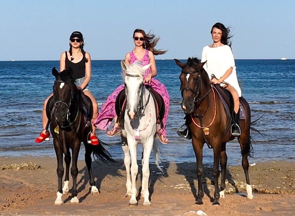 Three women ride horses along a sandy beach with the blue sea behind them; left rider wears black top and red sandals, center wears a pink dress, right wears a white dress.